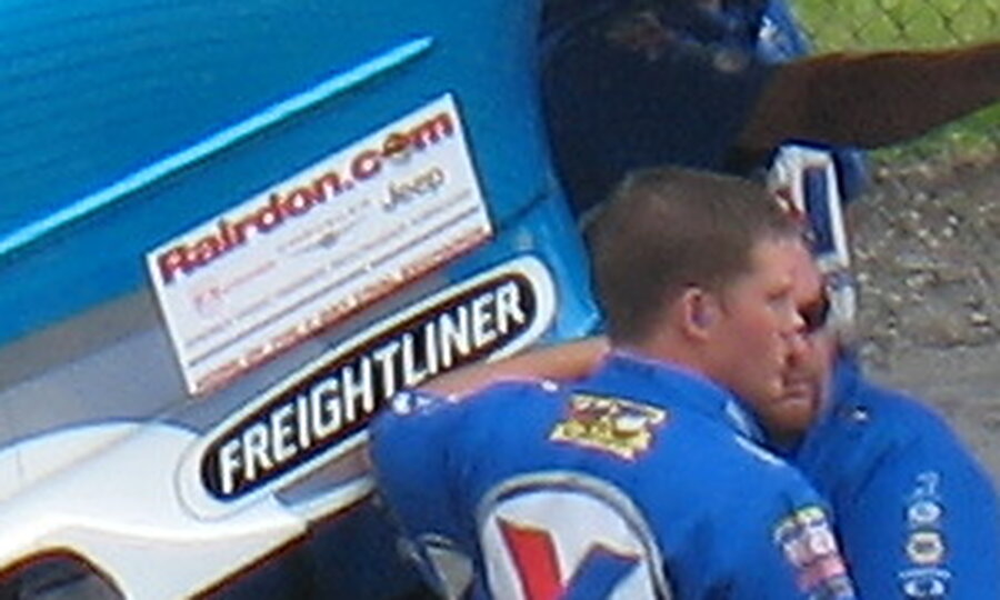 Close-up crop of the same Funny Car: a white Rairdon.com decal on the front fender, just behind the front wheel arch, directly above a black Freightliner decal, with a crew member in a blue shirt in front